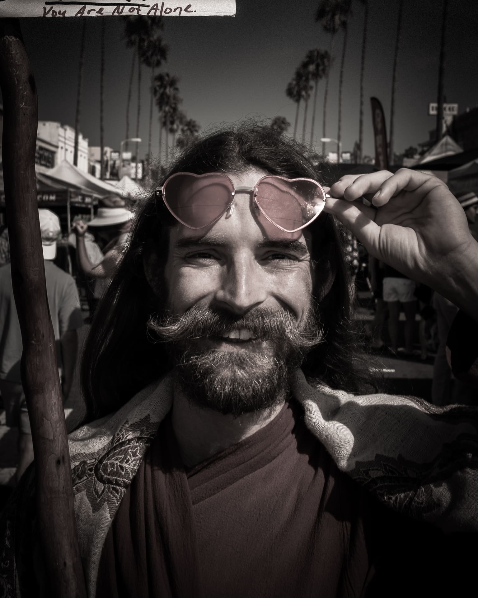 A smiling man with a mustache lifts pink heart-shaped sunglasses on a sunny street