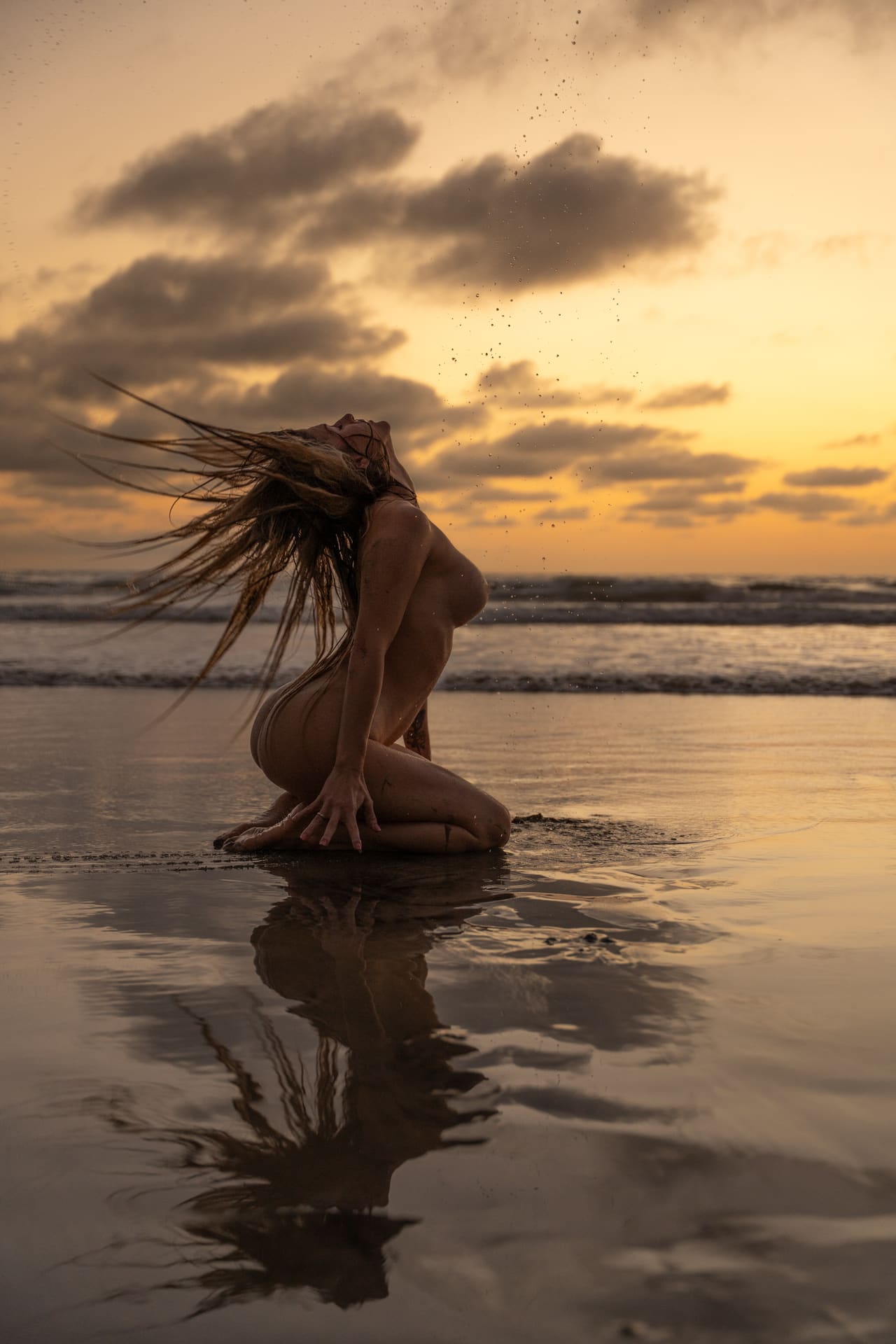 A nude woman kneels on wet beach sand at sunset, whipping her hair back