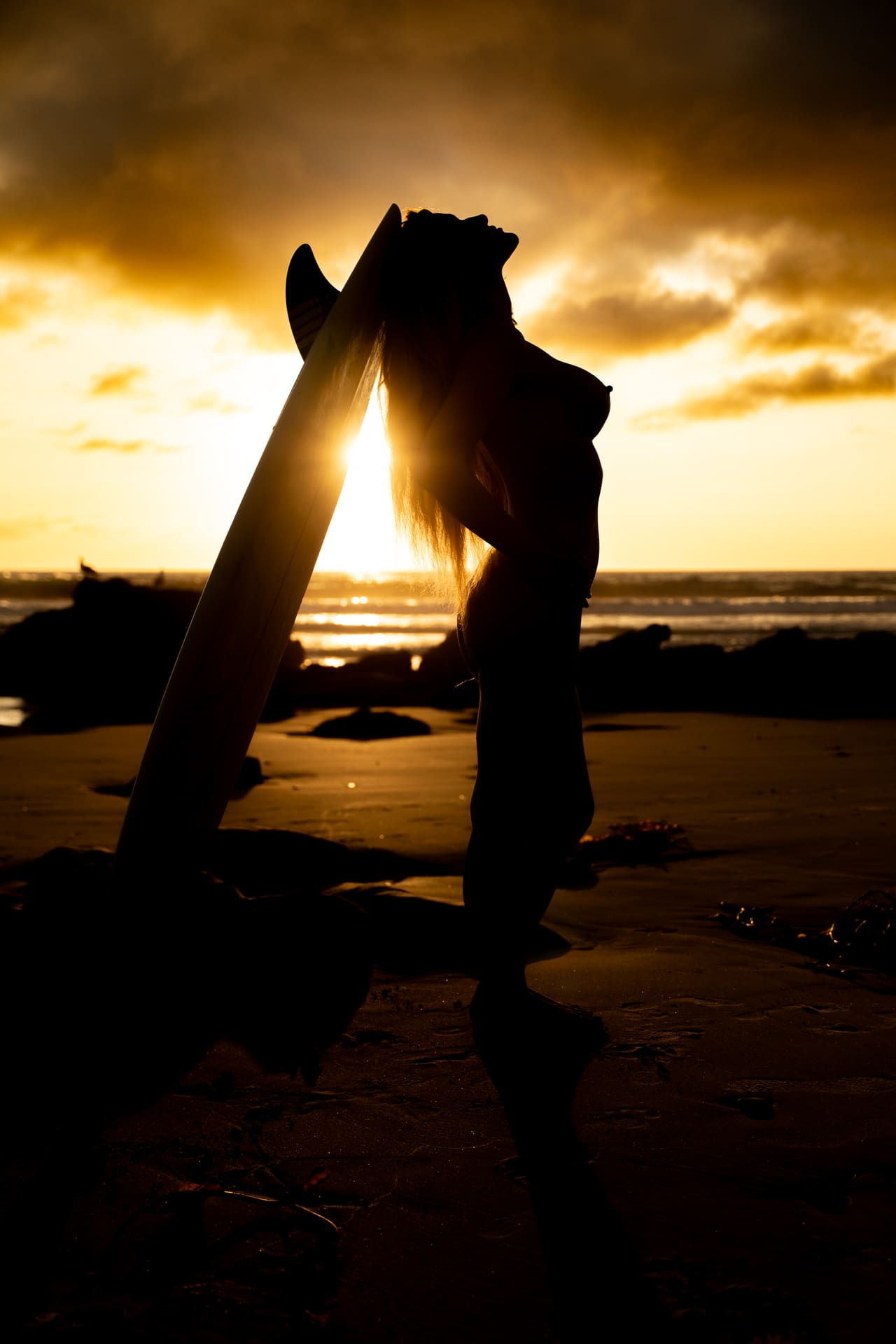 A silhouetted woman leans back against a surfboard on a beach at sunset