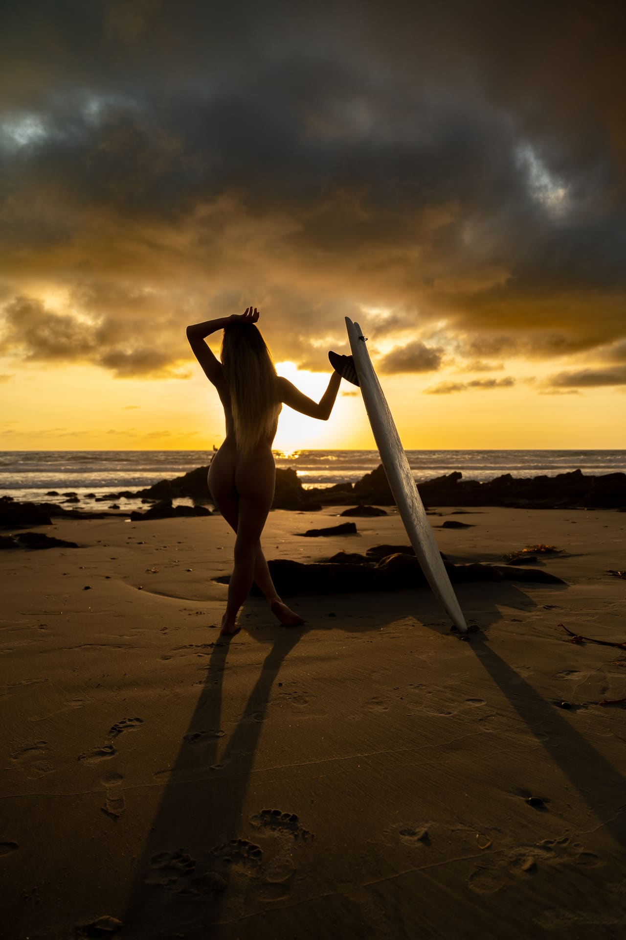 A silhouetted woman poses on a sandy beach beside a surfboard at sunset