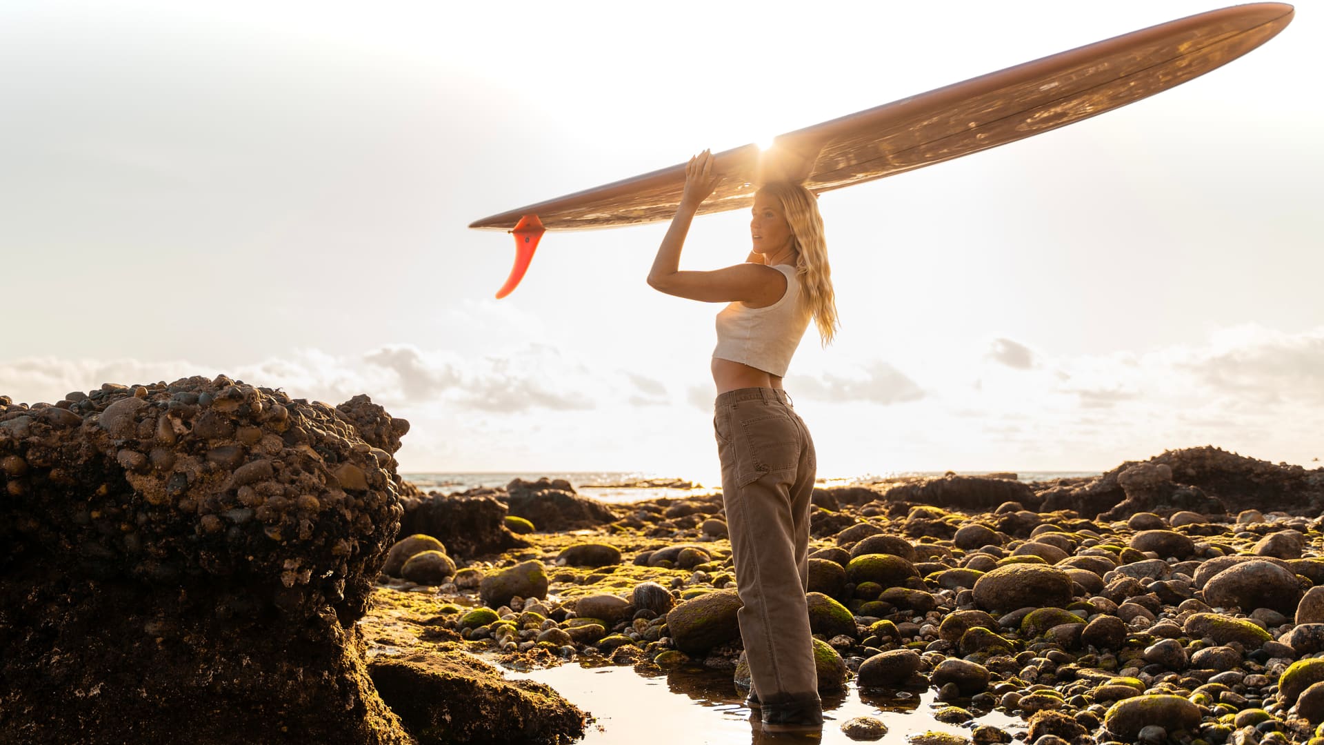 A woman carrying a surfboard on her head along a rocky shore at sunset