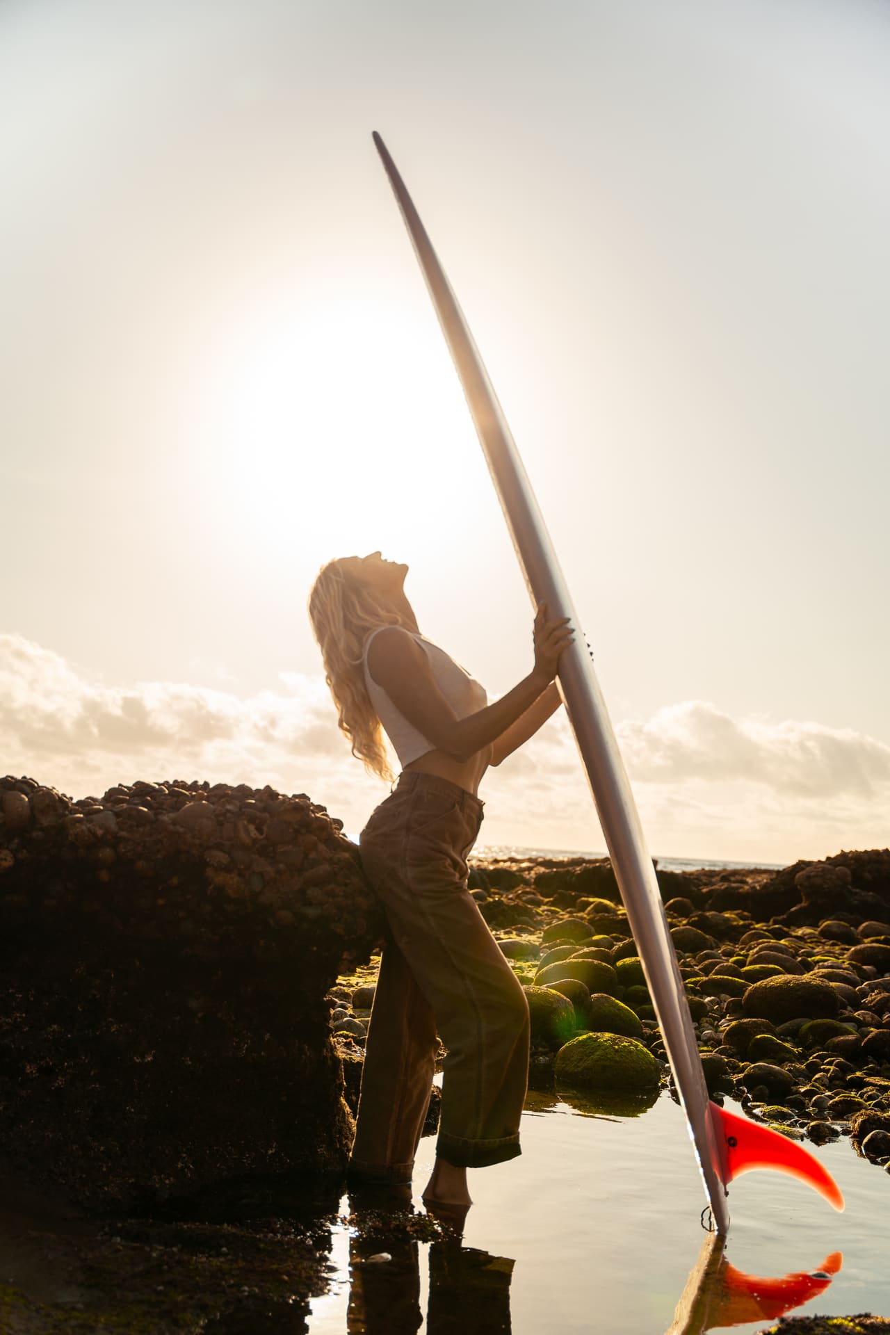 Woman holding surfboard upright on rocky shoreline during golden hour sunset