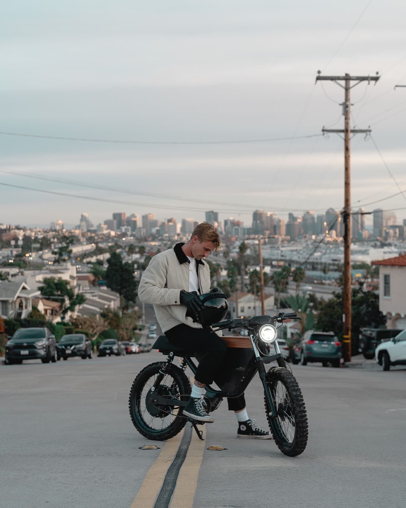 A man holding a helmet sits on an electric motorcycle overlooking a city skyline