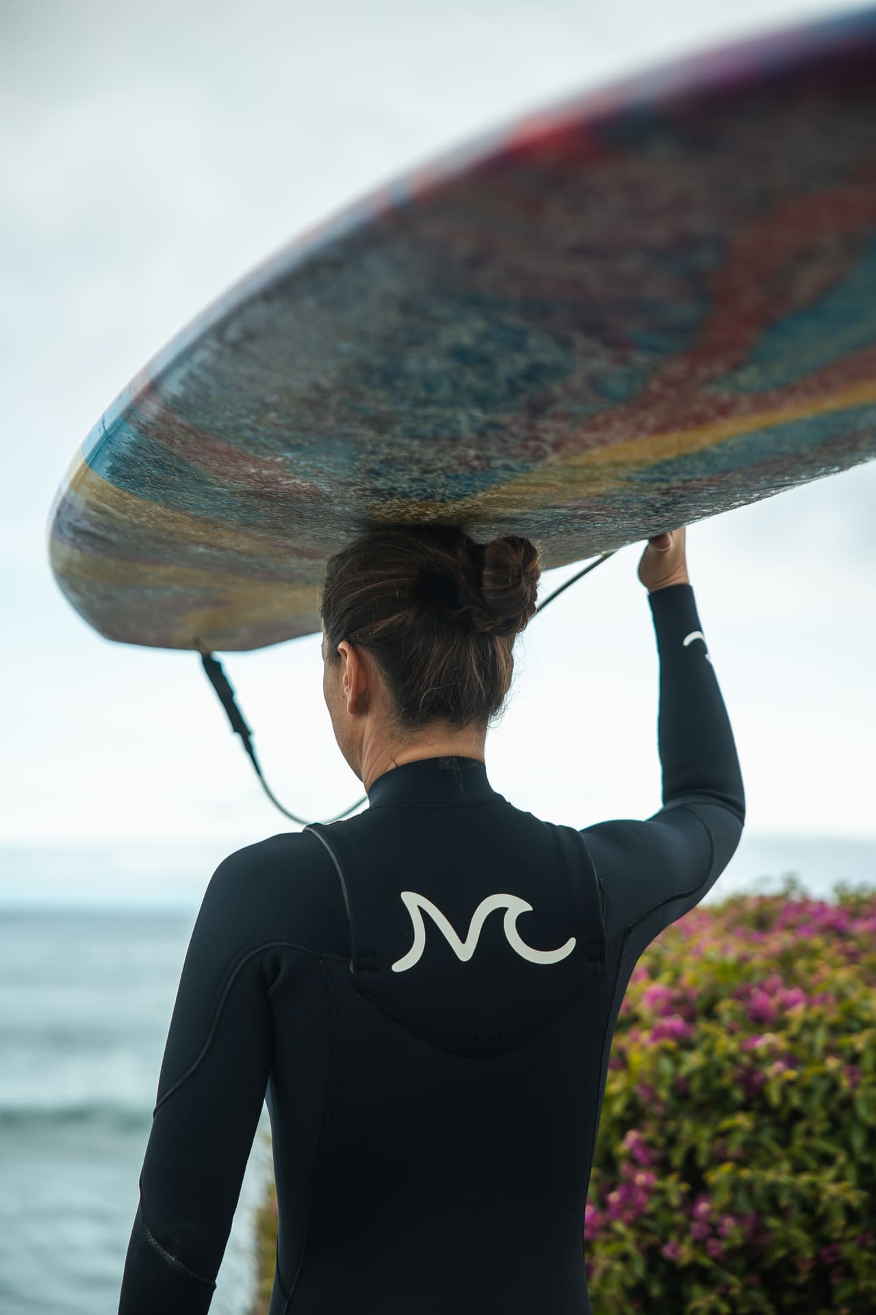 Surfer carrying colorful board overhead near ocean wearing black wetsuit
