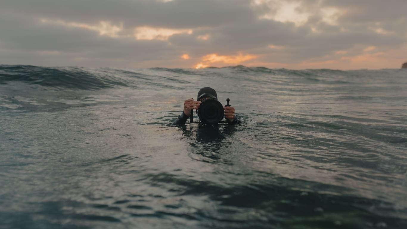 A photographer in a wetsuit holds an underwater camera housing in ocean waves at sunset