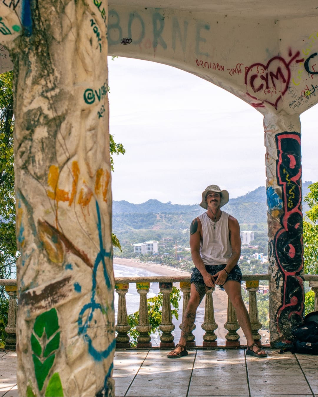A man sitting in a graffiti-covered gazebo overlooking a beach and distant mountains