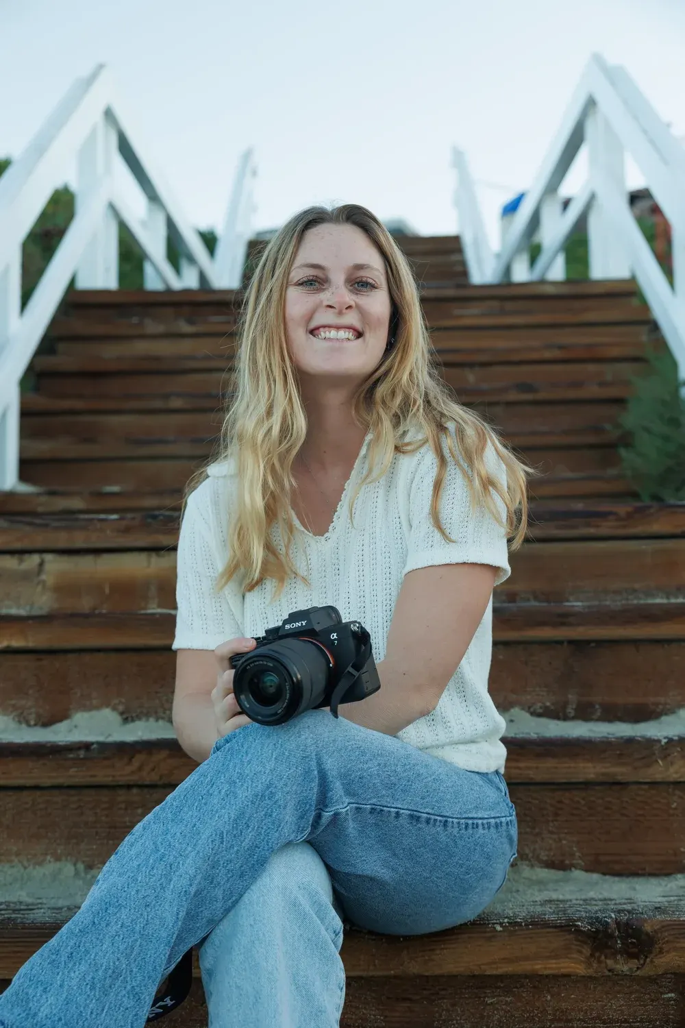 A smiling woman holding a black camera while sitting on wooden outdoor stairs