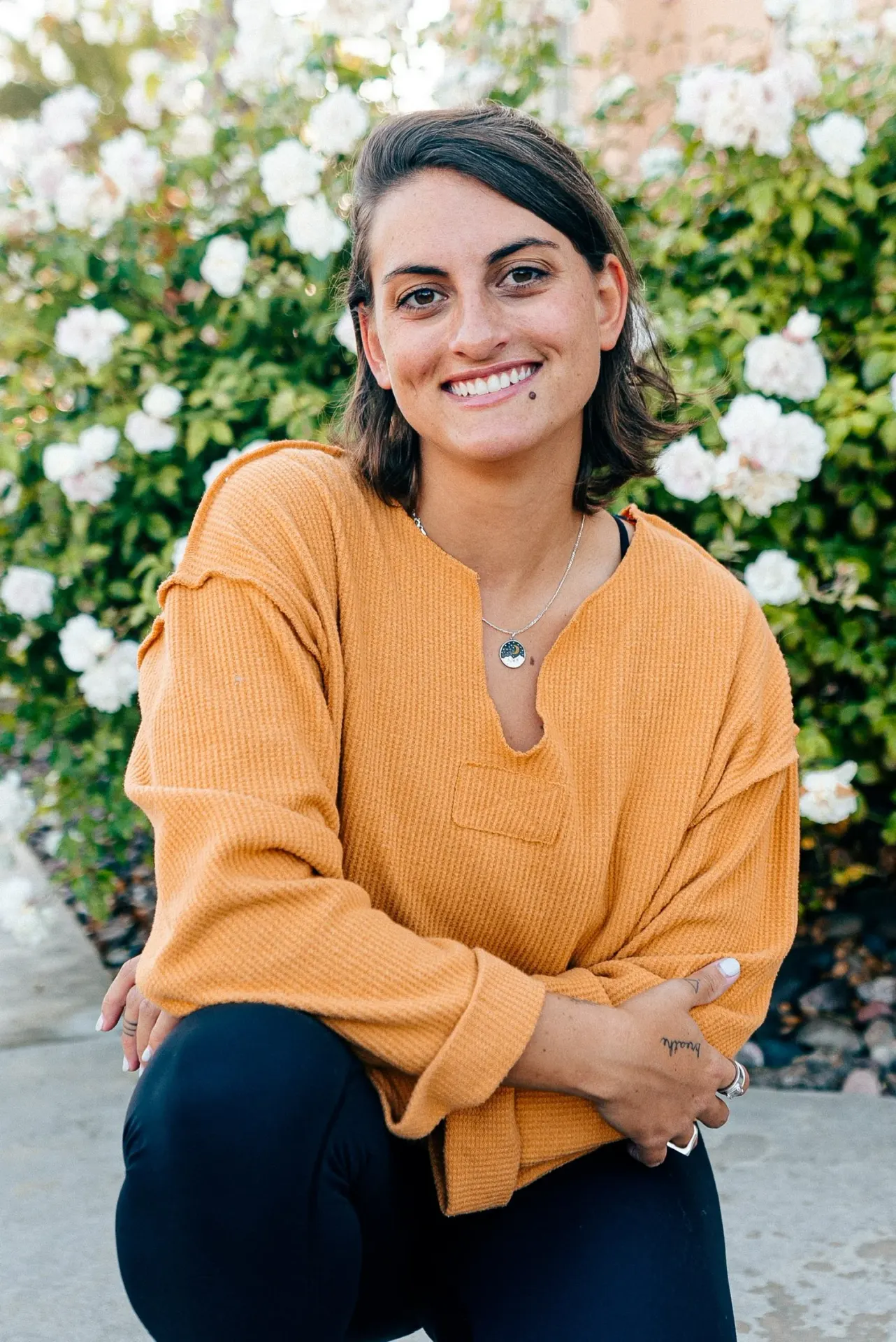 Smiling woman wearing a mustard yellow sweater poses outdoors in front of white rose bushes