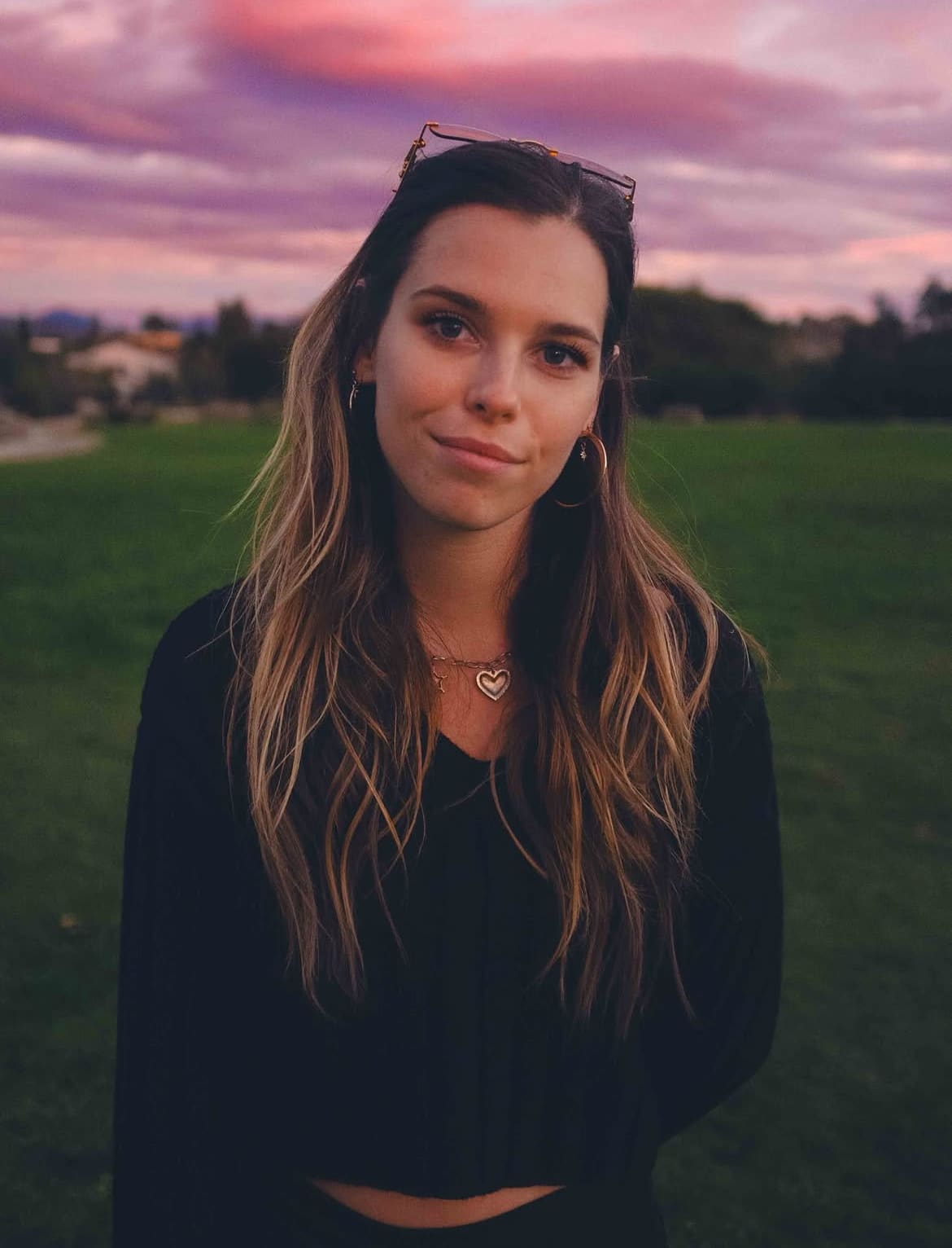 A young woman smiling in a grassy field against a pink sunset sky