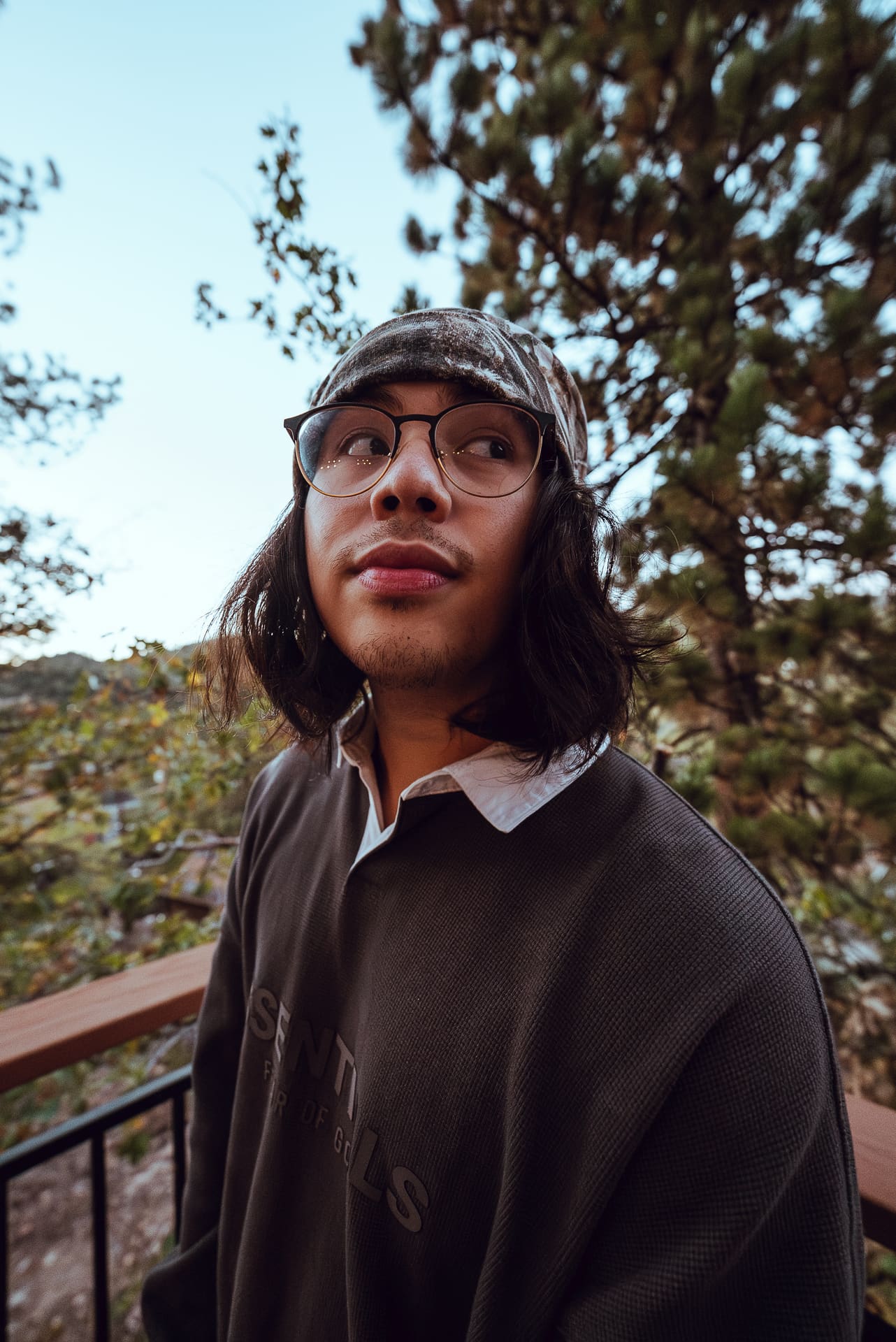 A young man wearing glasses and a patterned beanie looks upward outdoors among pine trees