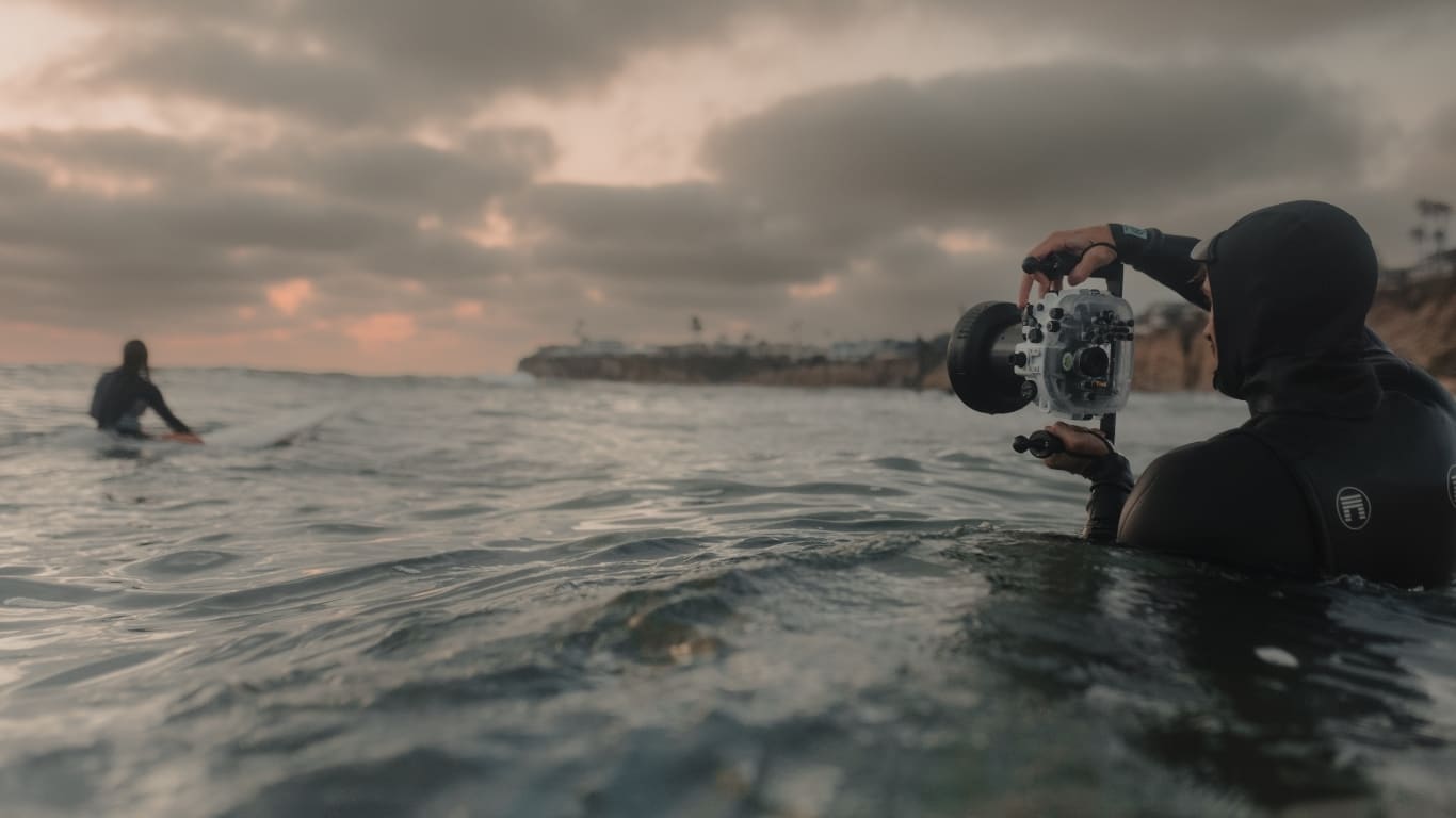 A photographer in a wetsuit holds a camera housing in the ocean facing a surfer