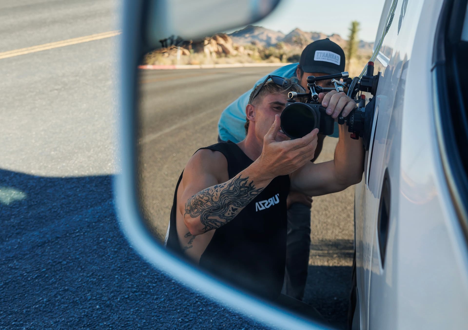 Side mirror reflection of a tattooed man adjusting a camera mounted on a white vehicle