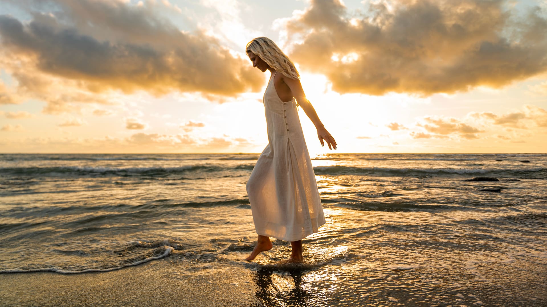Blonde woman in white linen dress walking barefoot along ocean shore at sunset