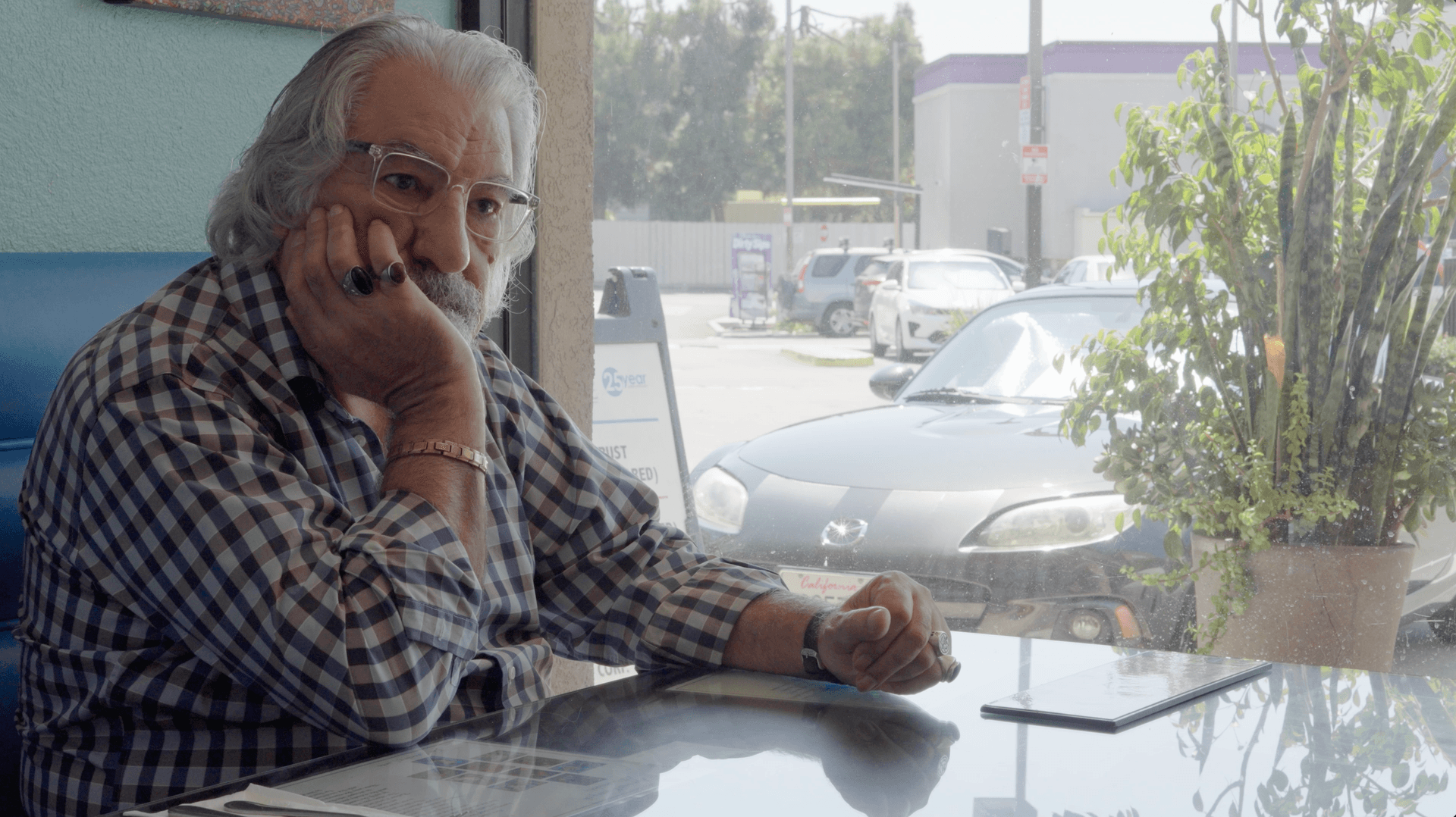 Older man with gray hair and glasses sits pensively at a window-side table