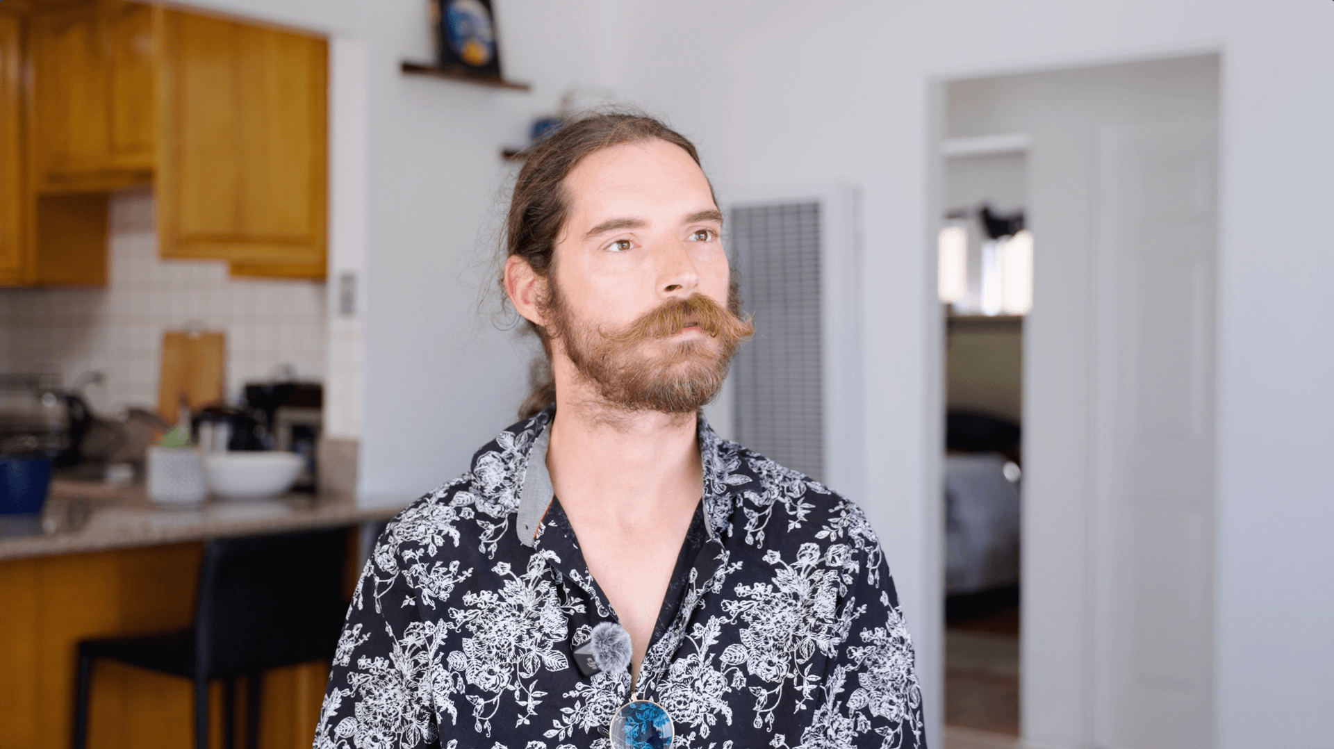 Bearded man with long hair in floral shirt standing indoors near kitchen