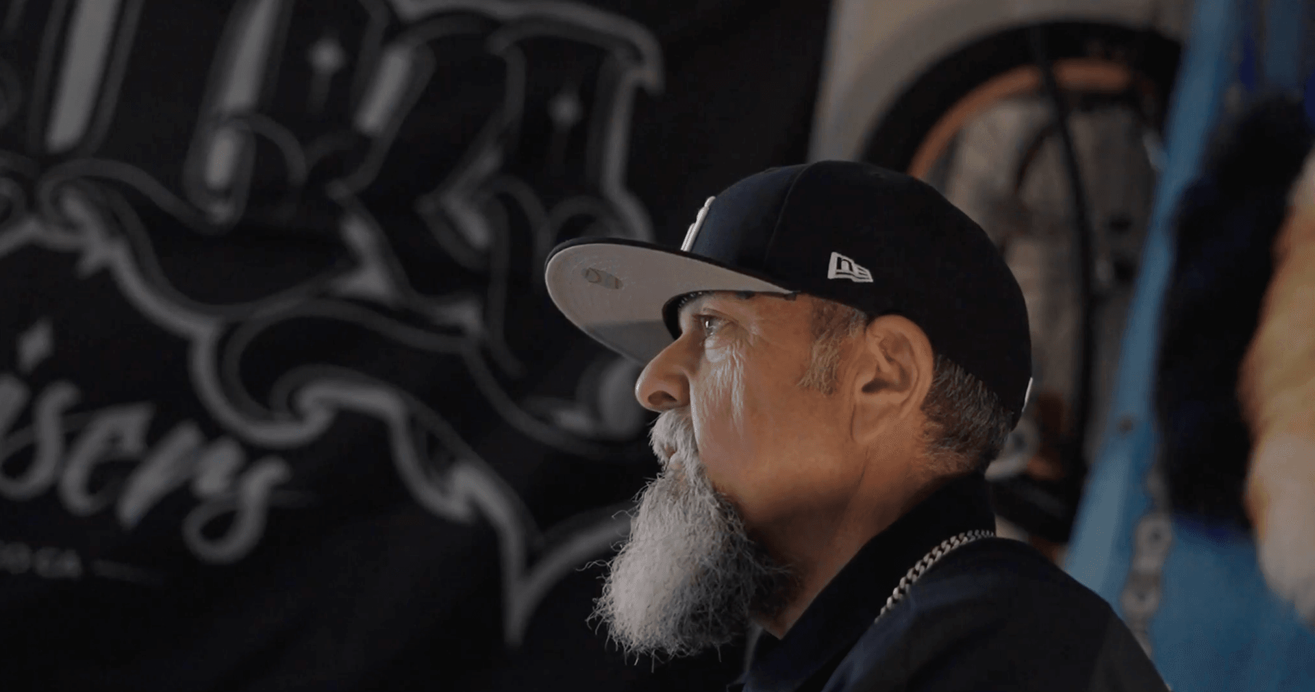 Older man with white beard and New Era cap gazes upward indoors