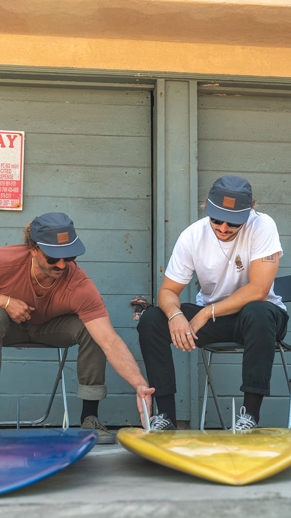 Two men sitting in chairs inspect a yellow surfboard lying on the ground