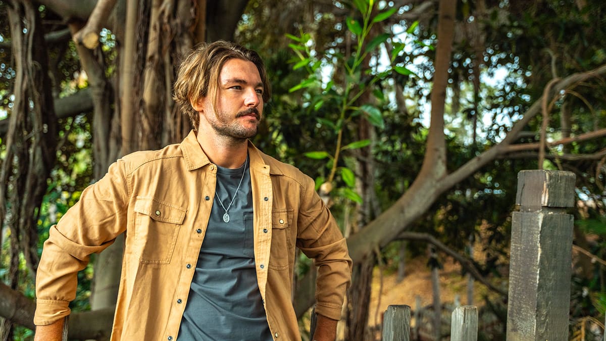 A man wearing an open tan shirt stands outdoors surrounded by trees