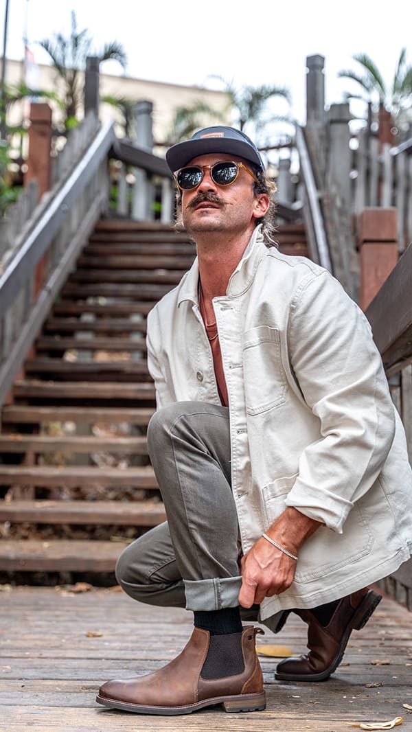 Man in sunglasses crouching on a wooden walkway adjusting his cuffed pant leg