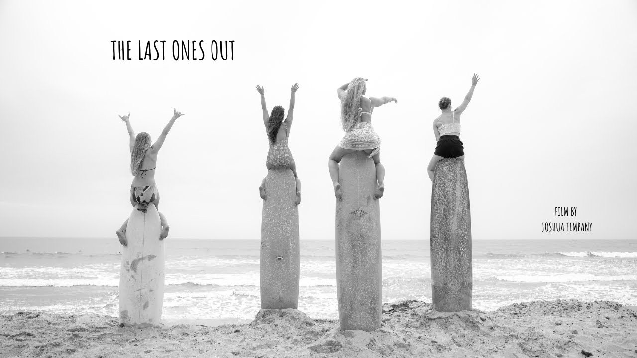 Four women sitting atop upright surfboards on a beach facing the ocean with raised arms