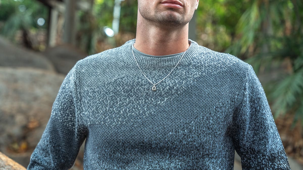 A man wearing a grey knit sweater and a silver pendant necklace outdoors