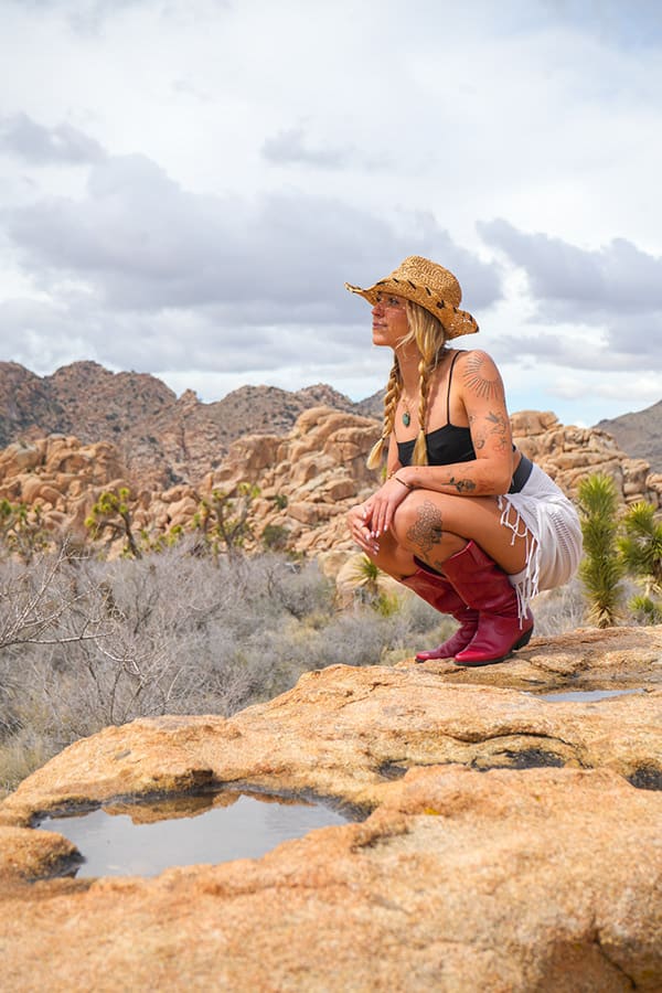 A woman in a straw hat and red boots crouches on desert rocks