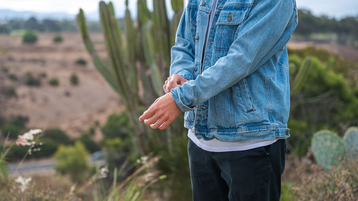 A man in a denim jacket adjusts his cuff in a desert landscape with cacti