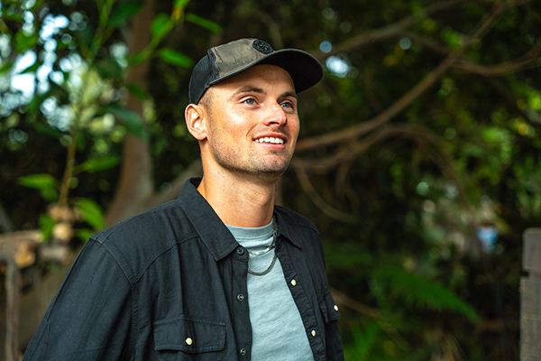 A smiling man wearing a dark cap and shirt standing outdoors with green foliage behind him