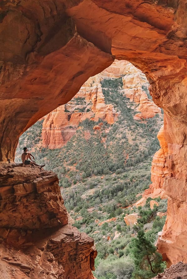 A man sits on a ledge inside a red rock arch overlooking a canyon