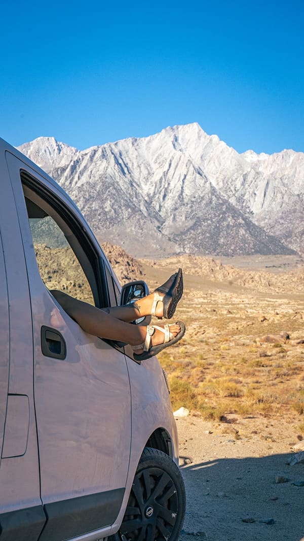Bare legs wearing sandals sticking out of a van window in front of desert mountains