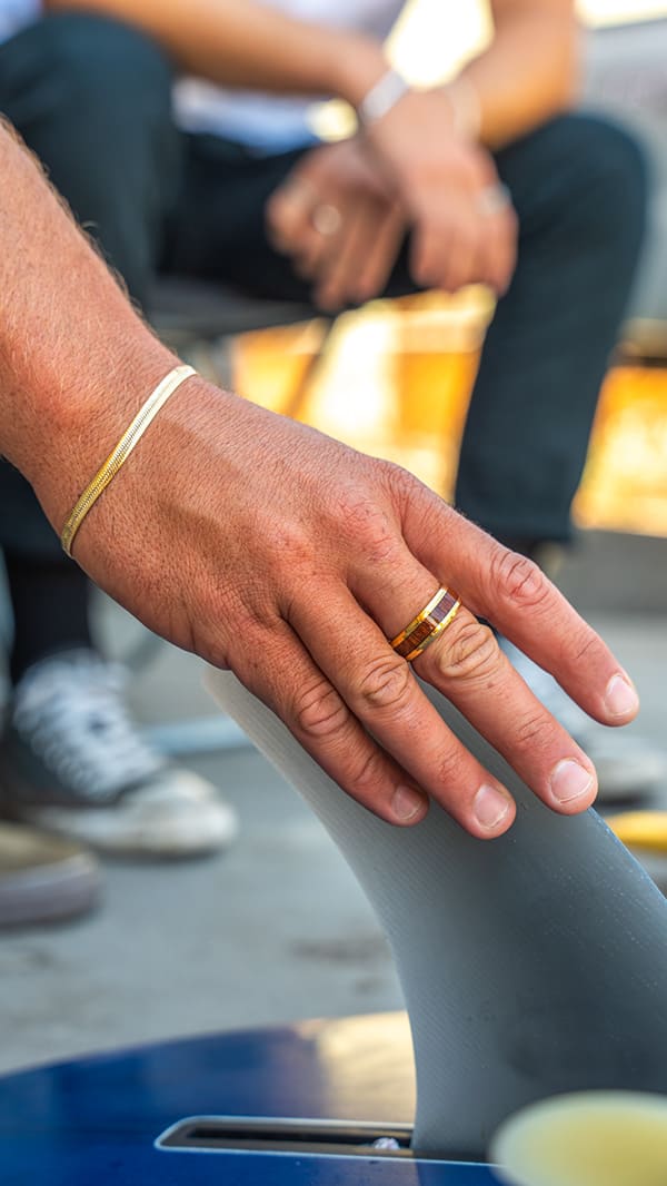 A tanned hand wearing a gold bracelet and ring rests on a surfboard fin