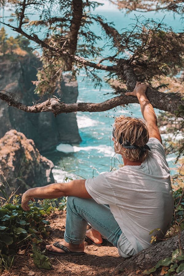 A man sits on a cliff under tree branches, looking out at the ocean
