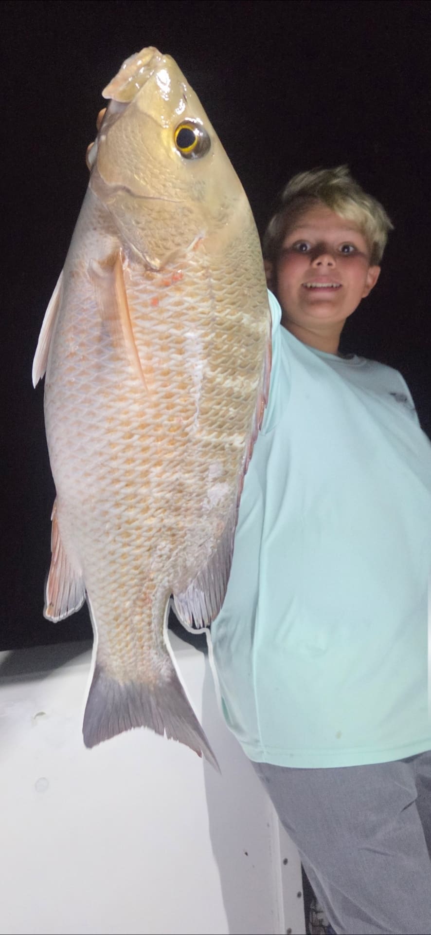 A young boy holding up a large snapper fish on a boat at night
