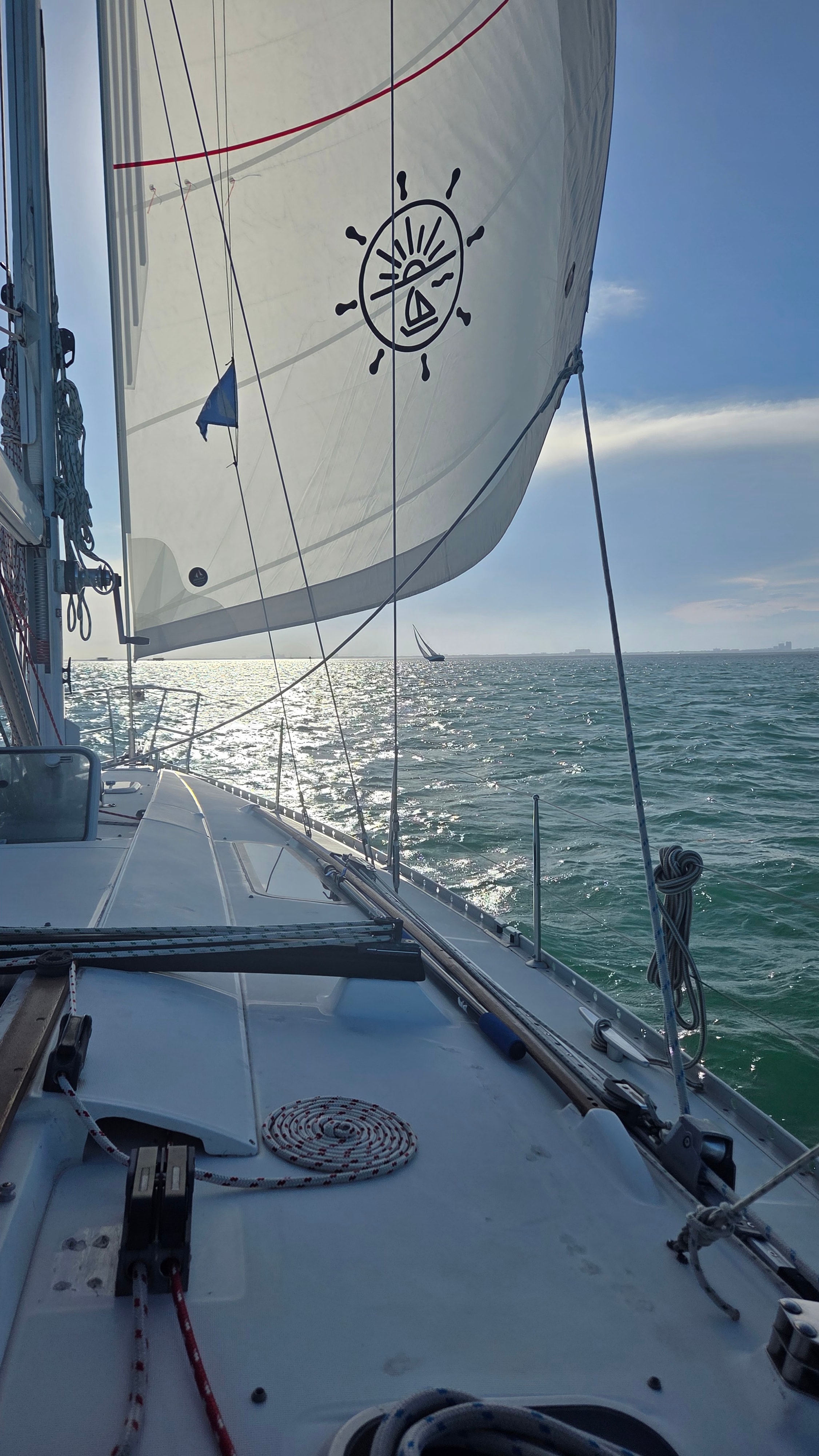 Sailboat deck view with white sail featuring nautical wheel logo on open sea