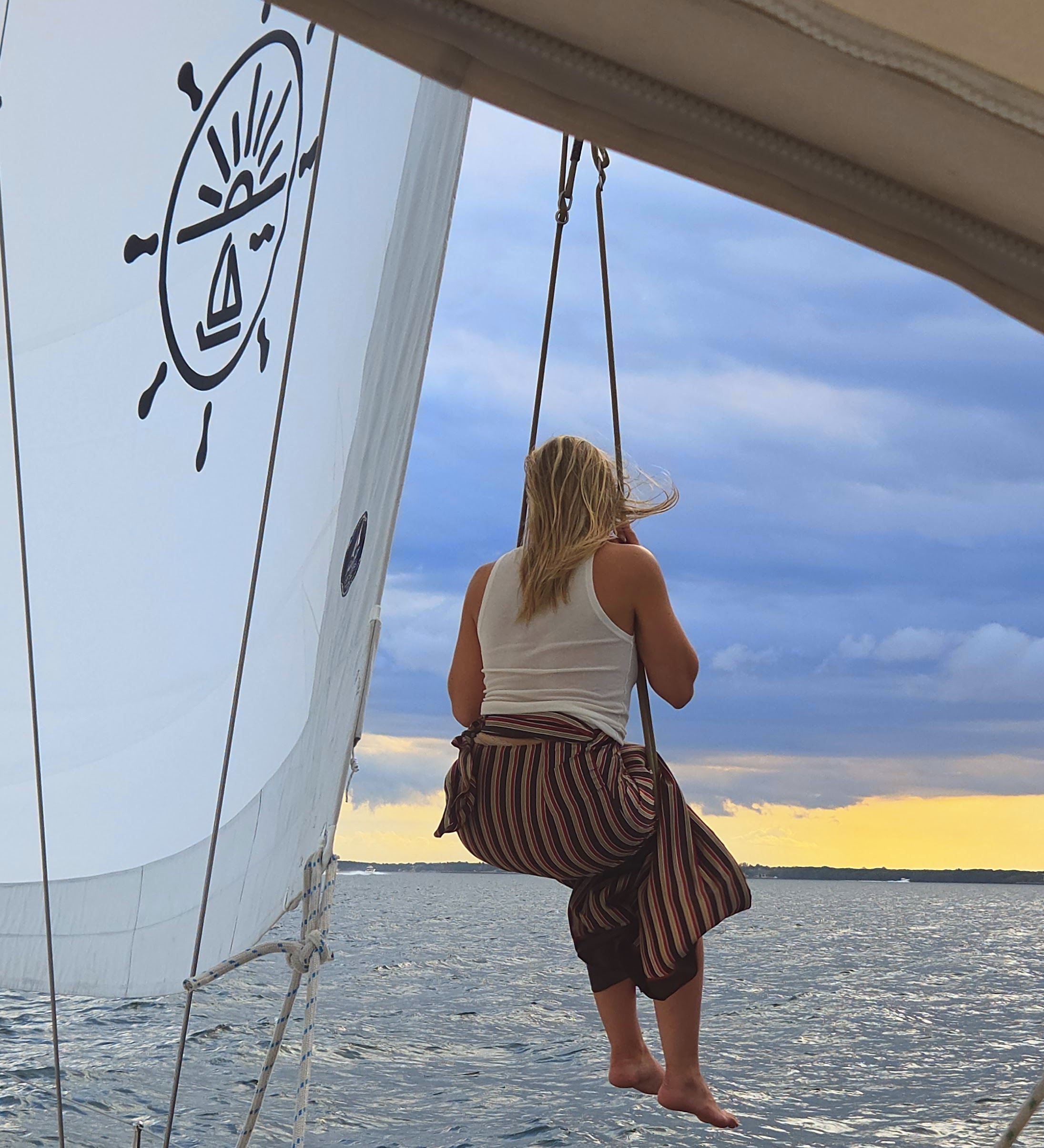 Barefoot woman sitting on rope swing hanging from sailboat over open water at sunset