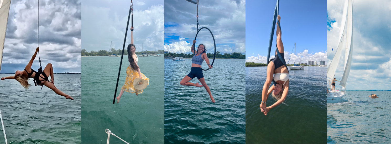 Women performing aerial acrobatics suspended from a sailboat mast over turquoise water