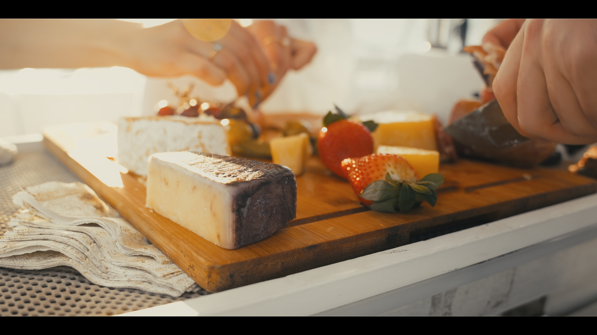 Hands preparing a charcuterie board with cheese wedges and fresh strawberries in bright sunlight