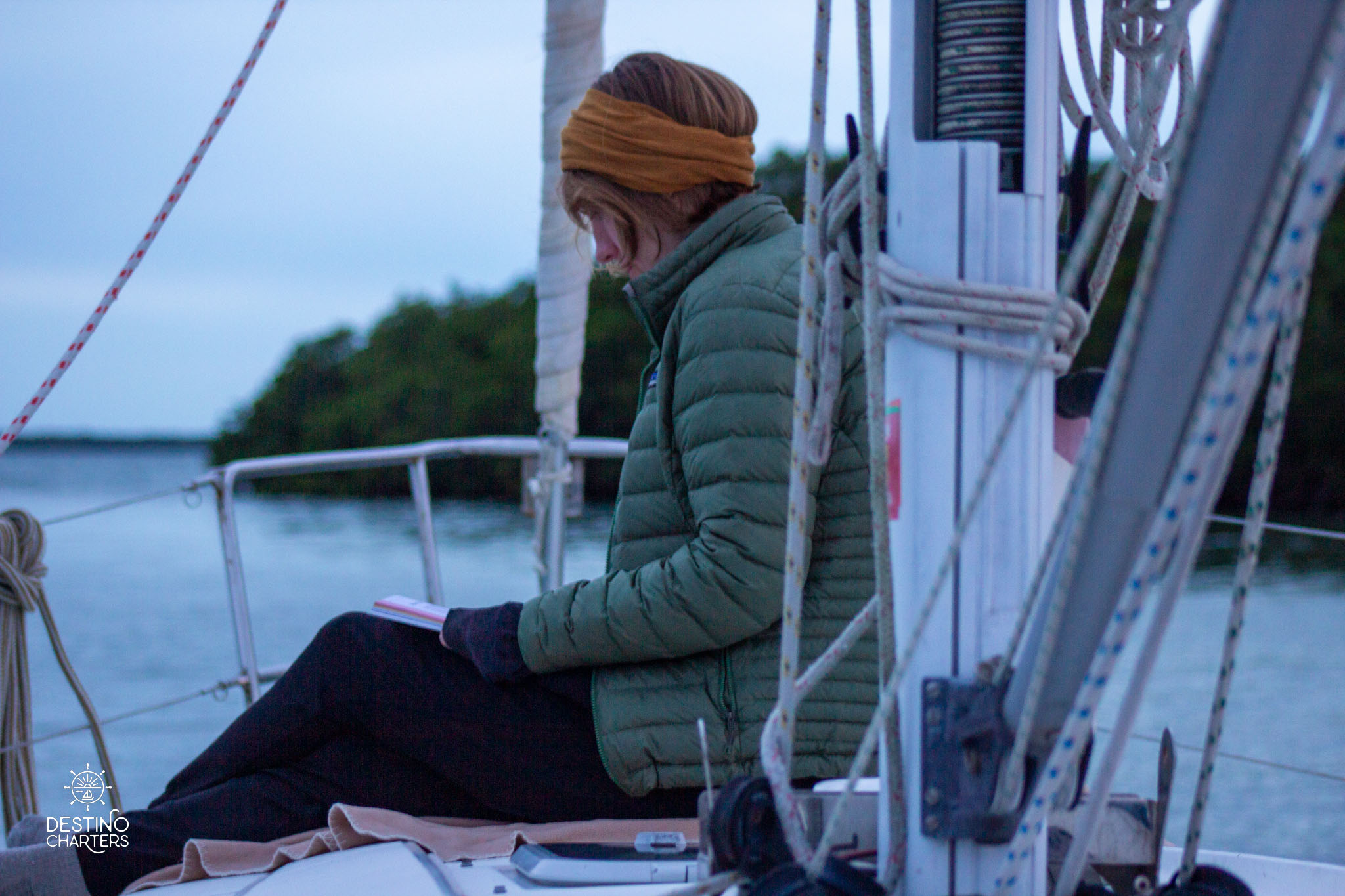 Woman reading on sailboat deck at dusk wearing green puffer jacket and headband