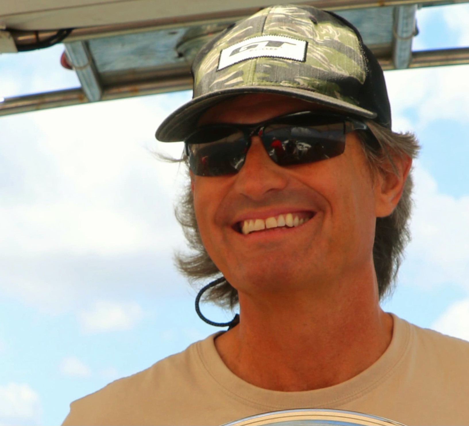 Smiling man in camo cap and sunglasses standing outdoors under blue sky