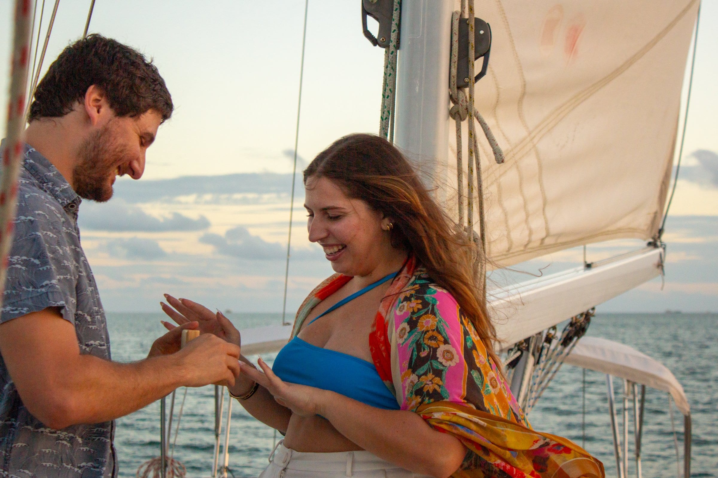 Man placing ring on woman's finger aboard a sailboat at sunset