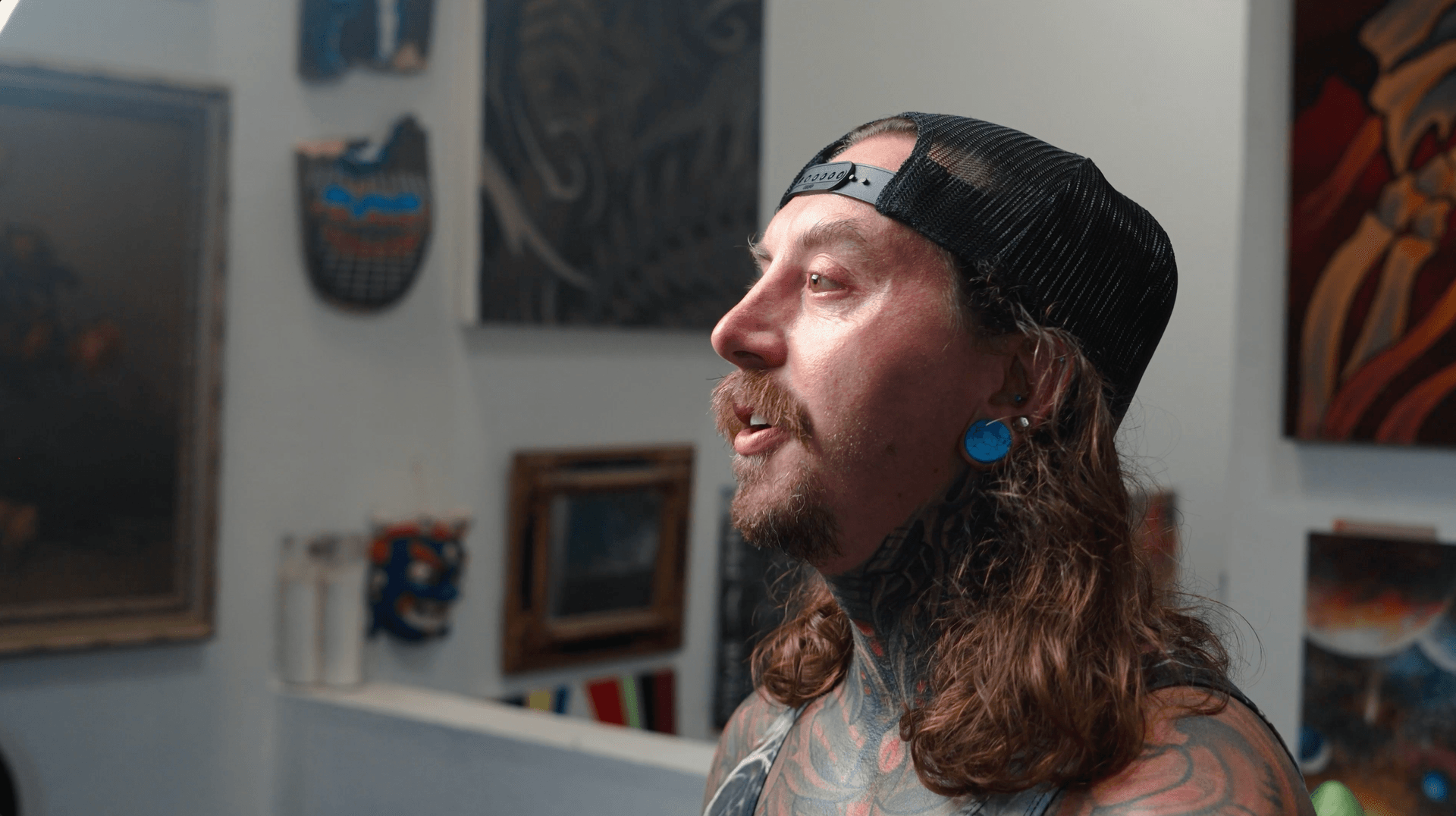 Tattooed man with long hair and ear gauges standing in an art-filled studio