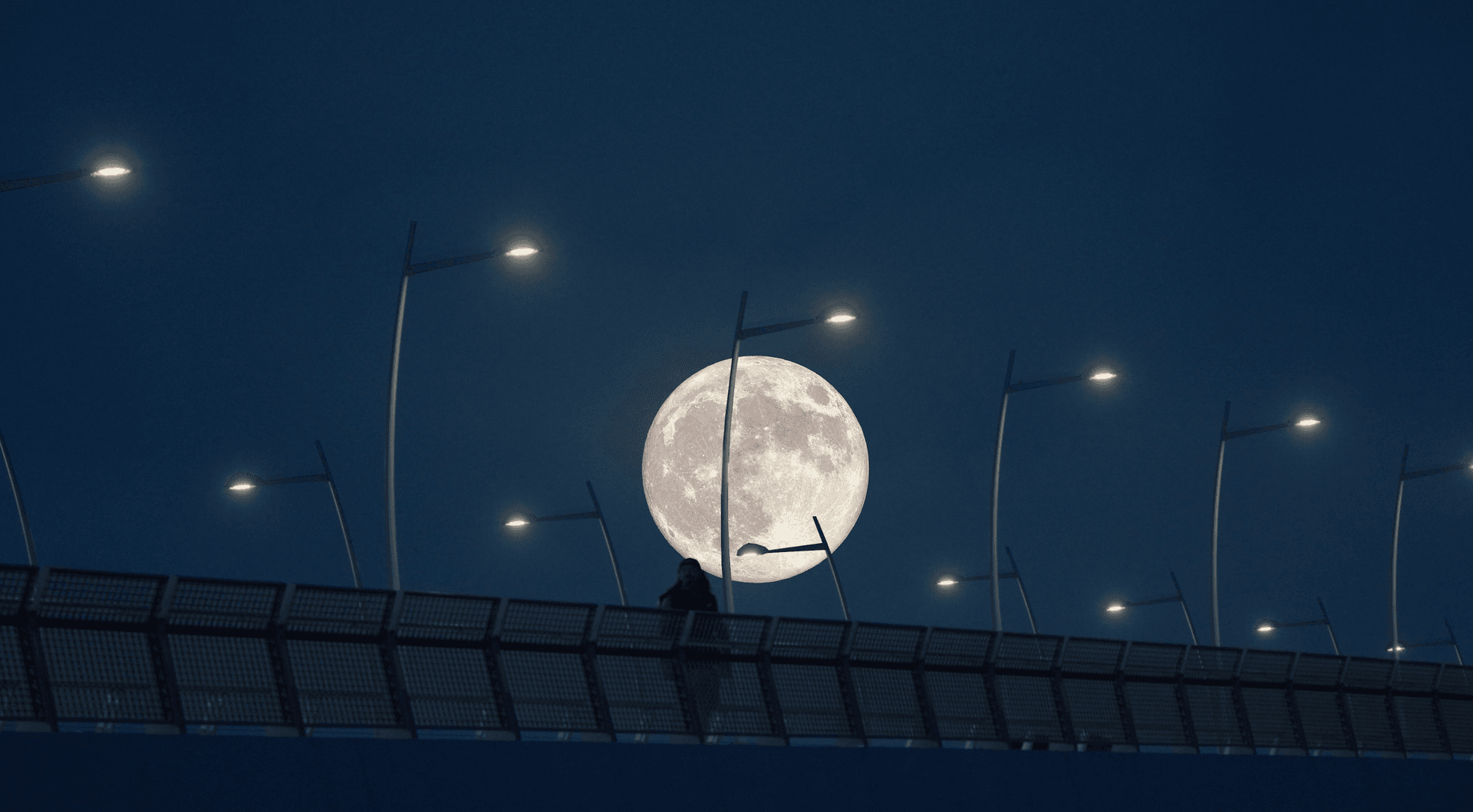 Full moon rising behind a pedestrian bridge with glowing street lamps at night