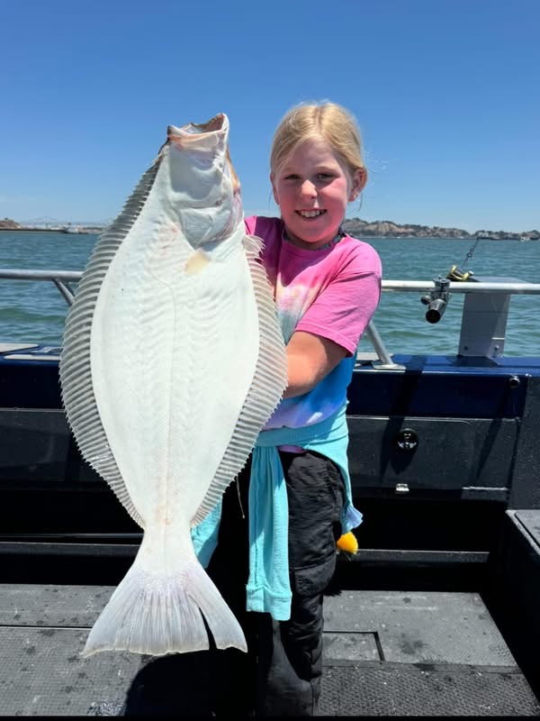 A young angler in a tie-dye shirt holds up a big California halibut on a Reel Addiction boat on San Francisco Bay