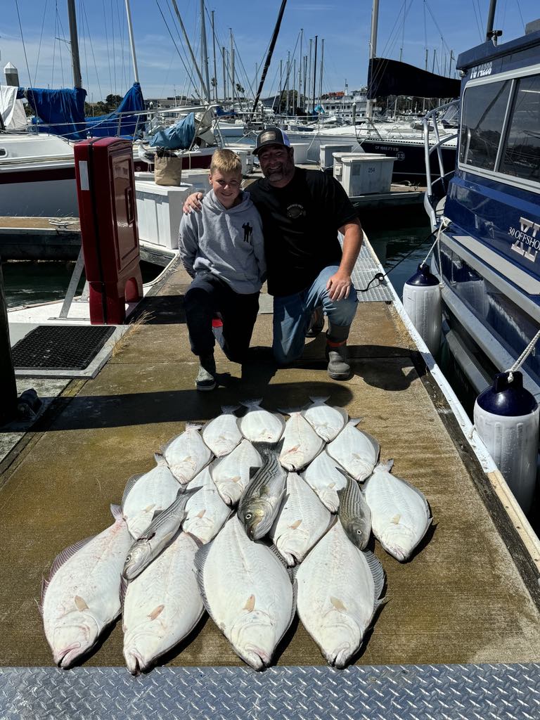 A father and son kneeling on the dock behind a pile of halibut and striped bass