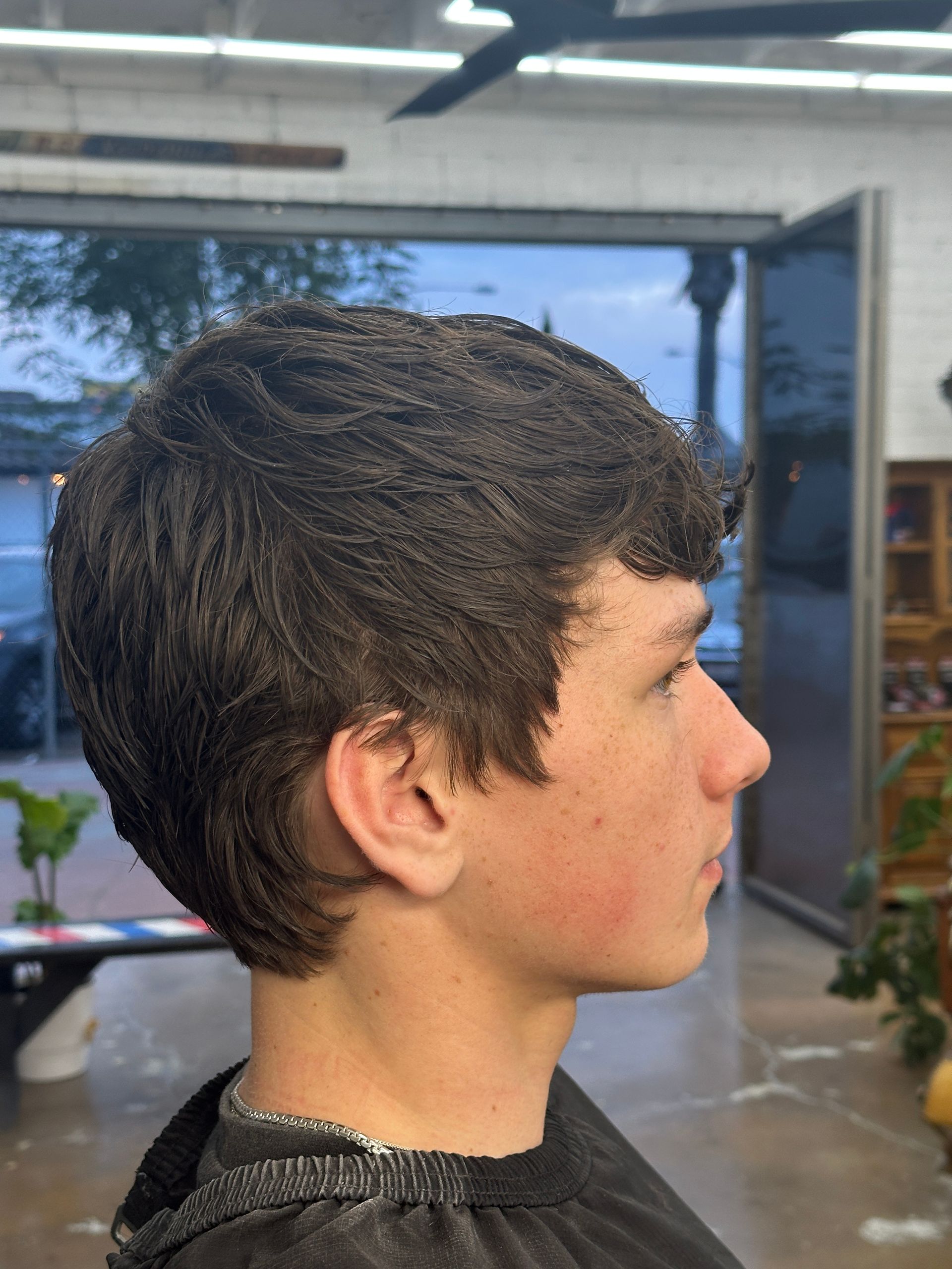 Young man with fresh layered haircut sits in barbershop wearing a black cape