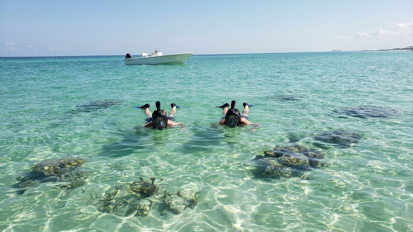 Two snorkelers in full-face masks floating over a shallow reef in clear turquoise water, a boat anchored behind them