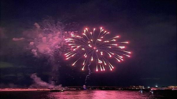 Pink fireworks bursting over the water at night, reflected in the bay, seen from the boat