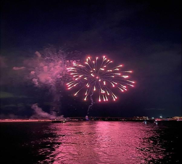 Pink fireworks bursting over the water at night, reflected in the bay, seen from the boat